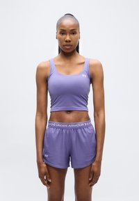 Γυναίκα που στέκεται στραμμένη προς τα εμπρός, φορώντας μοβ crop top και σορτς Under Armour, μπροστά σε απλό λευκό φόντο.