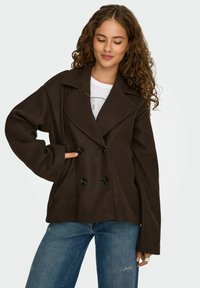 Bruine dubbelgeknoopte jas met brede revers, grote zwarte knopen en een relaxed fit. Gemaakt van zachte stof, met twee zijzakken.