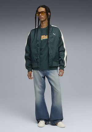 Grøn satinvindjakke med hvide striber, teal grafisk t-shirt, klokkeformede lyseblå jeans og hvide sneakers. Gule solbriller.