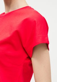 Κοντινό πλάνο ενός ατόμου που φορά ένα φωτεινό κόκκινο κοντομάνικο πουκάμισο με στρογγυλή λαιμόκοψη, πάνω σε απλό φόντο.