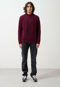 Maglione a maglia bordeaux con colletto alto, abbinato a jeans scuri e sneakers. Il maglione presenta una texture a costine e maniche a raglan.