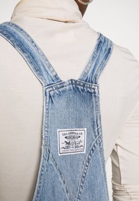 Ljusblå denimhängselbyxor med breda remmar, prydda med en vit Levi Strauss-etikett och synliga sömsdetaljer mot en beige topp.