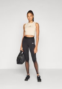 Svetlo béžový krátky tielko s logom, v kombinácii s čiernymi legínami s priehľadnými panelmi a bielymi pruhmi; drží čiernu športovú tašku.