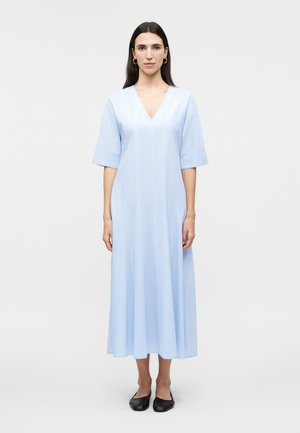 Γυναίκα που στέκεται με το σώμα στραμμένο μπροστά, φορώντας ένα ανοιχτό γαλάζιο midi φόρεμα με μανίκια μέχρι τον αγκώνα και μαύρα επίπεδα παπούτσια, μπροστά από ένα απλό φόντο.