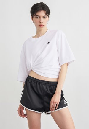 Junge Person mit kurzen dunklen Haaren, die ein weißes, geknotetes, bauchfreies T-Shirt und schwarze Sportshorts mit weißem Rand trägt.
