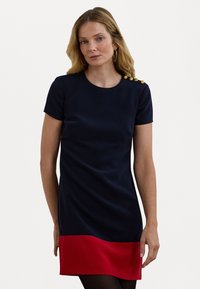 Vestido azul-marinho com uma bainha vermelha, mangas curtas e decote redondo. Apresenta botões dourados decorativos no ombro. Textura suave.