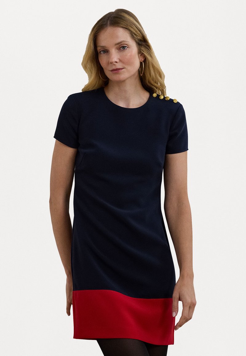 Vestido azul-marinho com uma bainha vermelha, mangas curtas e decote redondo. Apresenta botões dourados decorativos no ombro. Textura suave.