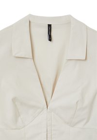 Blouse de couleur crème avec un col, un corsage structuré et des coutures de dart subtiles. Présente une patte de boutonnage centrale pour la fermeture.