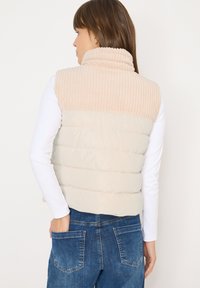 Chaleco acolchado beige con cuello acanalado y sección inferior lisa, usado sobre una camiseta blanca de manga larga, acompañado de jeans oscuros de mezclilla con bolsillos.