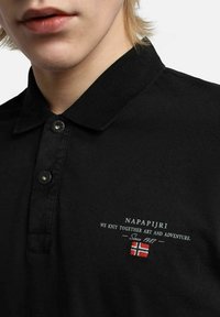 Musta polo-paita nappikauluksella, jossa on brodeerattu teksti "Napapijri" ja pieni Norjan lippu. Pehmeä, teksturoitu kangas.