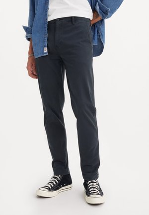 Personne portant un pantalon slim noir, des baskets montantes noires à lacets blancs, une chemise blanche et une veste en jean bleue avec les manches retroussées.