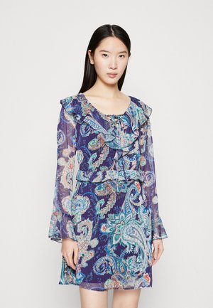 Ung kvinne iført en langermet lilla paisley-kjole med volanger, stående mot en ensfarget hvit bakgrunn.