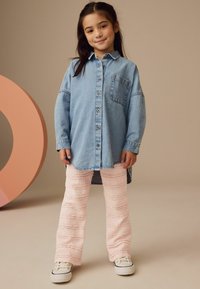 Denimhemd mit Tasche, Knopfverschluss und langen Ärmeln, kombiniert mit pinken, strukturierten Schlaghosen und weißen Turnschuhen.