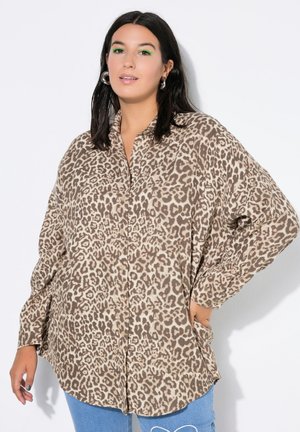 Donna con capelli scuri indossa una camicia beige e marrone con stampa leopardata oversize e jeans blu, posando con una mano in tasca.