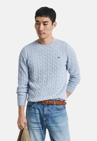 Helles blauer Kabelstrickpullover mit Rundhals und gerippten Bündchen. Trägt dazu eine hellblaue Jeans und einen braunen geflochtenen Ledergürtel.