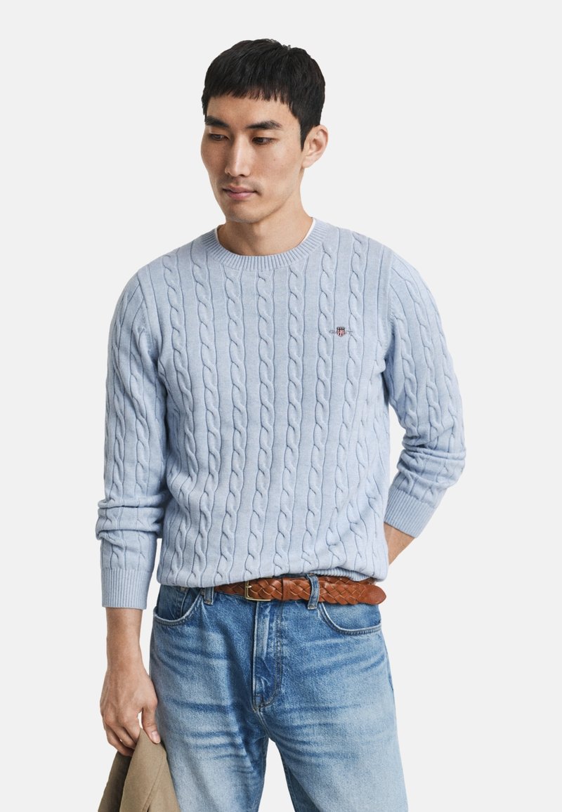 Helles blauer Kabelstrickpullover mit Rundhals und gerippten Bündchen. Trägt dazu eine hellblaue Jeans und einen braunen geflochtenen Ledergürtel.