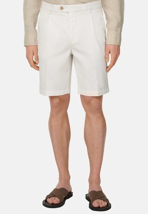 Homme portant un short blanc sur mesure, une chemise beige à manches longues et des sandales marron, debout devant un fond uni.