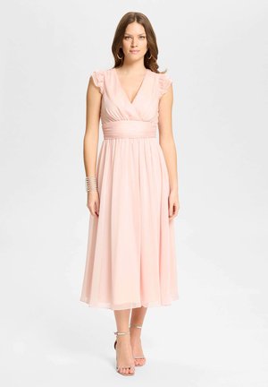 Mujer con un vestido midi sin mangas de color rosa claro con hombros con volantes, pendientes de aro plateados, pulsera y tacones con tira en el tobillo, de pie frente a un fondo blanco.