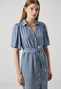 Vestido de camisa em denim com mangas curtas e volumosas, gola, botões à frente e um cinto amarrado na cintura. Cor azul clara.