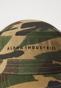 Tejido con patrón de camuflaje y texto bordado en negro "ALPHA INDUSTRIES" con detalles de costura visibles.