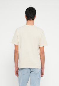 Lühikeste tumedate juustega mees, kes kannab lihtsat beežikat lühikeste varrukatega t-särki ja helerohelisi teksaseid, seisab seljaga valge tausta ees.