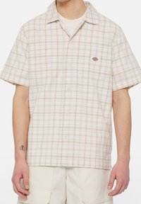 Uomo che indossa una camicia a scacchi beige a maniche corte sopra una t-shirt bianca, con pantaloni cargo beige.