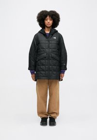 Czarna pikowana kurtka z kapturem, zapinana na zamek z przodu, z dwiema dużymi kieszeniami na piersi. Noszona na fioletowym swetrze i brązowych spodniach. Czarne buty.