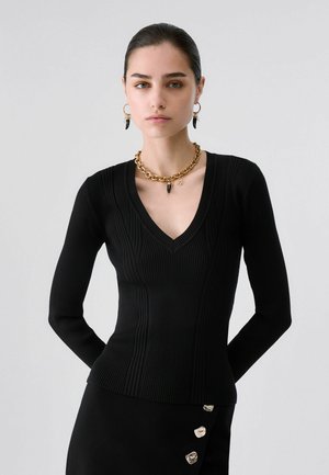 Frau mit dunklen Haaren, die einen schwarzen, gerippten V-Ausschnitt-Pullover, eine goldene Kette, passende Ohrringe und einen schwarzen Rock mit silbernen Knöpfen trägt.