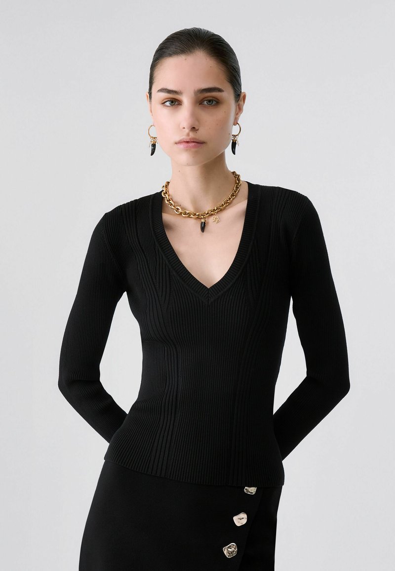 Femme aux cheveux foncés portant un pull noir côtelé à col en V, un collier chaîne doré, des boucles d'oreilles assorties et une jupe noire avec des boutons argentés.