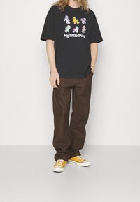 T-shirt en coton noir avec un graphique coloré "My Little Pony". Associé à un pantalon ample marron et des baskets jaunes. Coupe décontractée et design casual.
