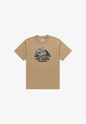 Beige katoenen T-shirt met een zwart grafisch ontwerp van een houthakker met een skateboard en bomen, met de tekst "DE DOERS" eronder.