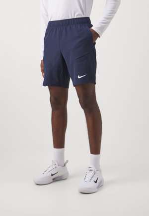 Mann trägt navyblaue Nike-Shorts mit Seitentaschen, weiße Sportschuhe und weiße Kniestrümpfe, steht vor einem einfarbigen Hintergrund.