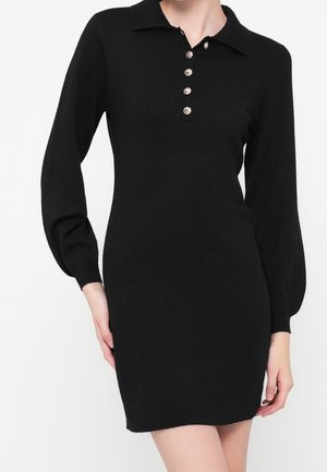 Woman wearing a fitted black knitted dress with long sleeves, a collar, and a front button closure with five silver buttons.