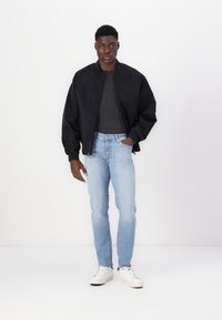 Chaqueta bomber negra con puños acanalados, camiseta gris oscuro, vaqueros de mezclilla azul claro y zapatillas blancas con detalles negros, de pie sobre un fondo blanco.