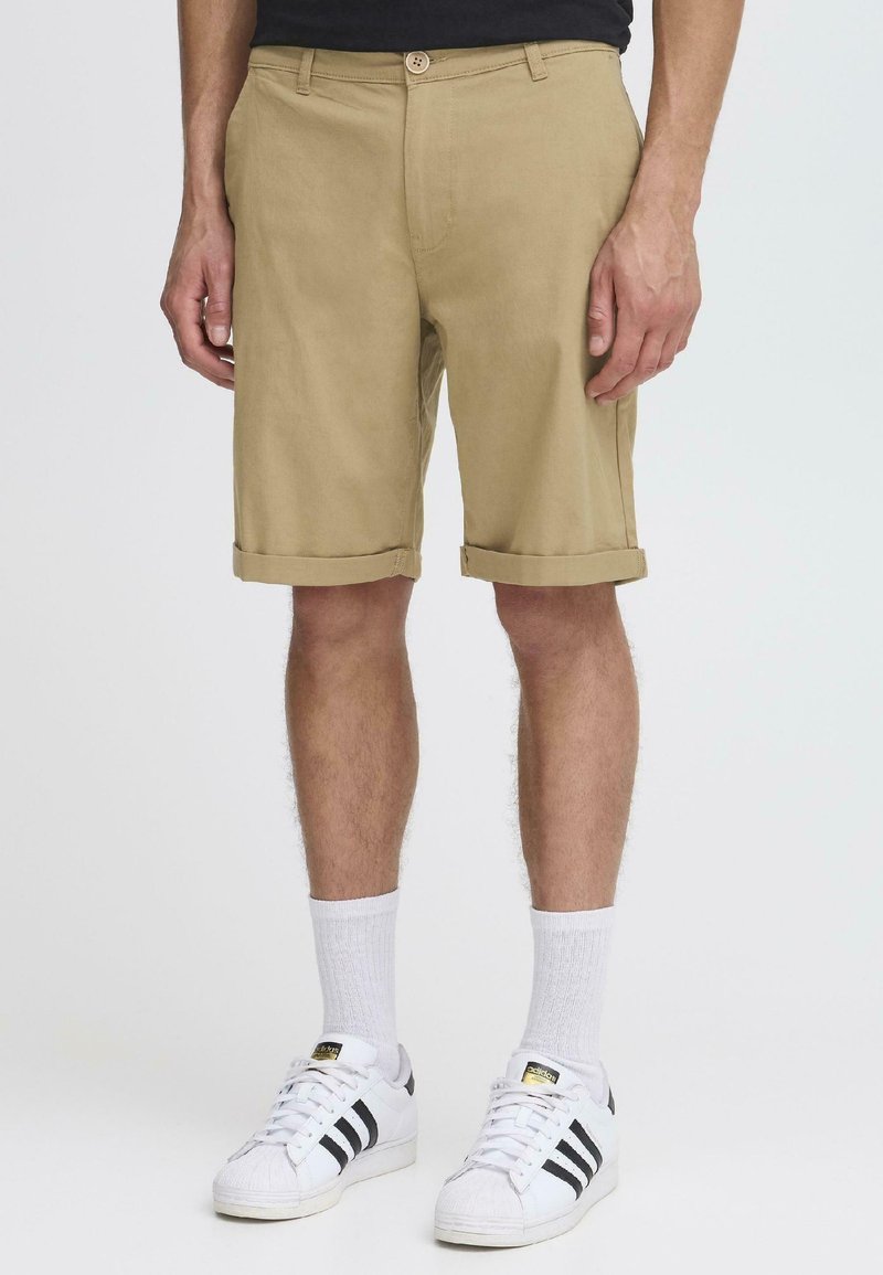 Pantalones cortos beige de algodón, con dobladillo enrollado, bolsillos frontales y cierre de un solo botón, combinados con calcetines blancos y zapatillas.