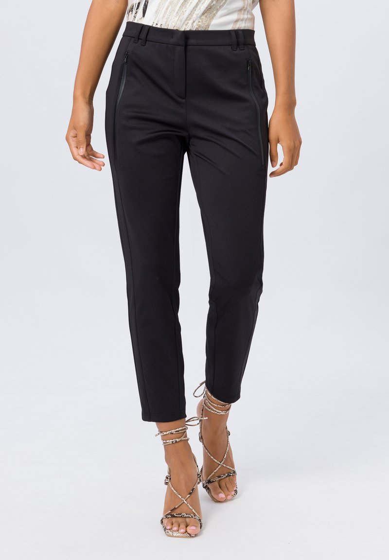 Un pantalon noir ajusté présente un design cintré, des poches zippées et une texture lisse. Associé à des sandales métalliques à brides.