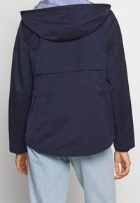 Dunkelblaue Kapuzenjacke mit einem geschichteten Rückendesign. Ausgestattet mit einer glatten Textur und einem abgerundeten Saum. Akzentuiert mit genähten Nähten.