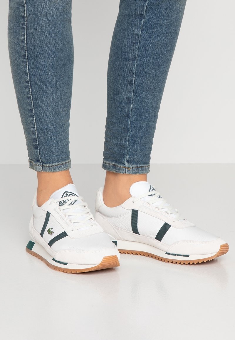 Baskets blanches de sport avec des bandes vertes, fermeture à lacets et semelle extérieure en caoutchouc texturée. Présentent un logo et des accents contrastants au talon.