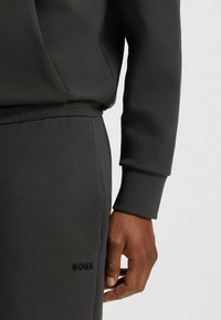 Completo sportivo grigio scuro composto da una felpa con cappuccio e zip, dotata di tasca a marsupio, e pantaloni joggers abbinati con il logo "BOSS" in rilievo sulla gamba.