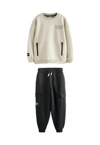 Felpa beige chiaro con due tasche con zip e logo; abbinata a pantaloni jogger neri con due tasche laterali e vita con cordoncino.