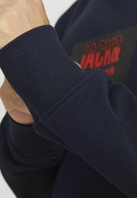 Navy sweatshirt manchet met ribbelstructuur, voorzien van een zwarte vierkante patch met rode tekst. Close-up toont de stikseldetails en een hand die het vastgrijpt.