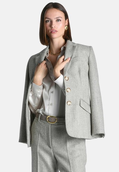 Femme aux cheveux bruns raides portant un blazer chevron gris clair drapé sur les épaules, un pantalon assorti, un chemisier en satin, un collier chaîne dorée et des boucles d'oreilles.