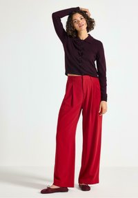 Kvinna med lockigt hår som bär en mörk lila knäppt cardigan, vida röda byxor och vinröda ballerinas, poserar med en hand på huvudet.