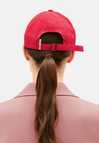 Red baseball cap with a rear adjustable strap. Cotton material, rounded crown, and a visible brand tag on the strap. Long brown ponytail visible.