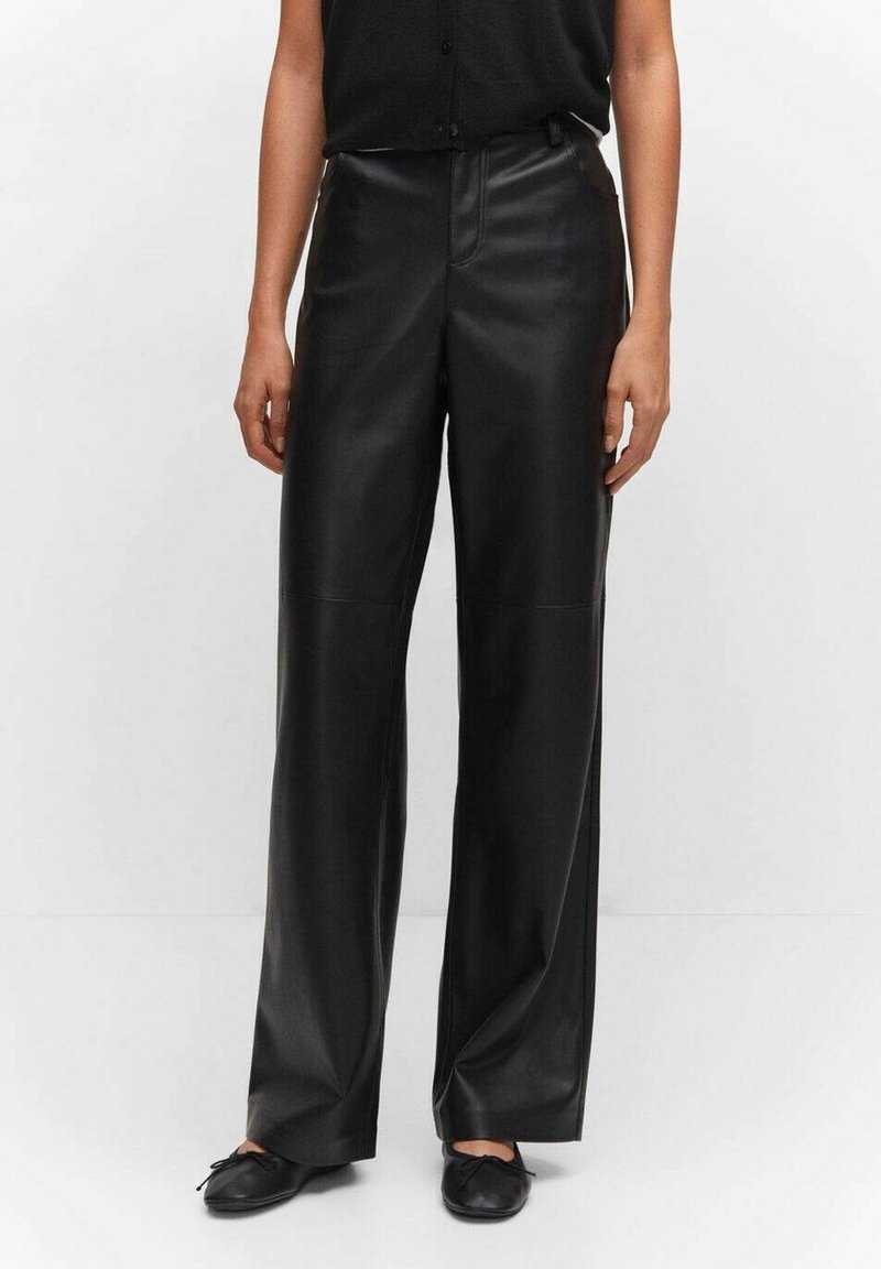 Personne portant un pantalon en cuir noir à jambes larges, un haut sans manches noir et des ballerines noires, debout devant un fond uni.