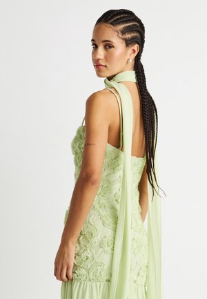 Γυναίκα με μακριά πλεγμένα μαλλιά, φορώντας ένα ανοιχτό πράσινο φόρεμα με υφή λουλουδιών και μια μακριά κασκόλ δεμένη γύρω από το λαιμό της, κοιτάζοντας πάνω από τον ώμο της.