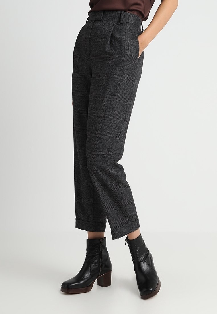 Personne portant un pantalon gris foncé sur mesure longueur cheville avec revers et des bottines en cuir noir à talons, debout avec une main dans la poche.