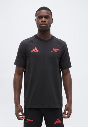 Mężczyzna stoi ubrany w czarną odzież sportową z czerwonym działem klubu piłkarskiego Arsenal i logo Adidas na koszulce i szortach.