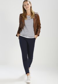 Brun læderjakke, stribet langærmet top, navy bukser og hvide sneakers. Modellen står selvsikkert mod en lys baggrund.
