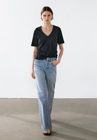 Kvinde, der går fremad iført en sort V-hals T-shirt, lyseblå brede jeans og sorte sandaler foran en ensfarvet hvid væg.